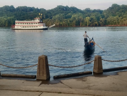 A gondola glides on calm water near a riverboat, surrounded by lush greenery and a peaceful shoreline.