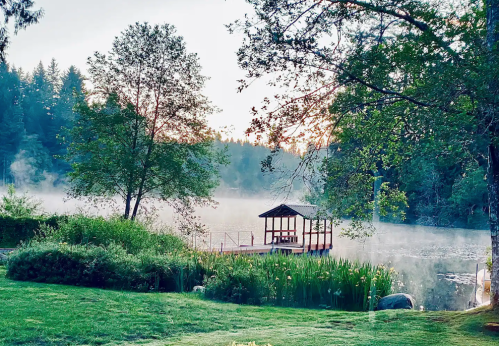 A serene lakeside scene at dawn, featuring a misty lake, lush greenery, and a small wooden dock.