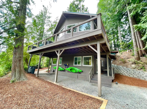 A modern house surrounded by trees, featuring a deck and kayaks on the side, set in a natural landscape.