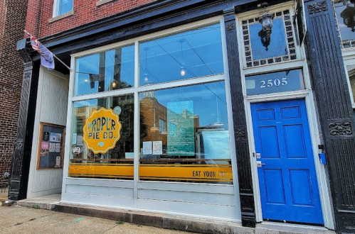 Exterior of Proper Pie Co. with large windows, a blue door, and a sign that says "Eat Your Pie."