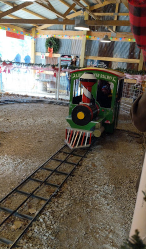 A small, festive train on a circular track inside a decorated barn, surrounded by holiday decorations.
