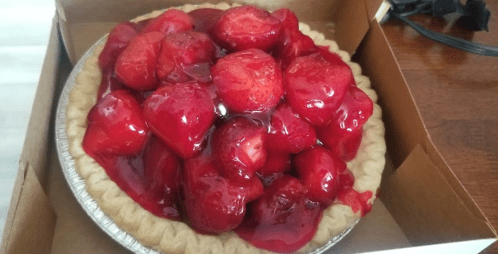 A freshly baked strawberry pie with a glossy red glaze, sitting in a cardboard box.