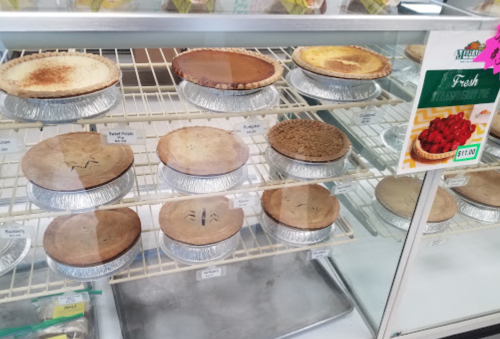 A display case filled with various pies on metal trays, labeled with names and prices.