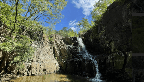 A serene waterfall cascades down rocky cliffs, surrounded by lush greenery and a clear blue sky.