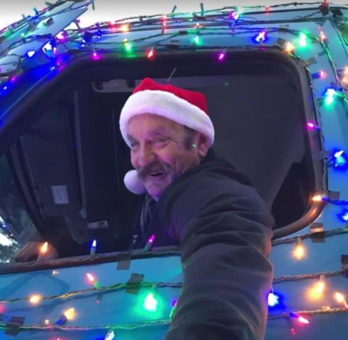 A smiling man in a Santa hat leans out of a decorated truck adorned with colorful Christmas lights.