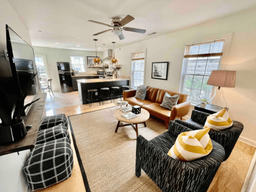 Cozy living room with a brown sofa, patterned chairs, a coffee table, and a kitchen in the background. Bright and inviting space.