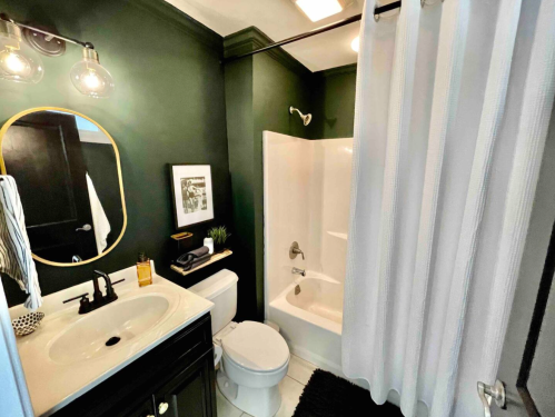 A modern bathroom featuring dark green walls, a white shower curtain, and a sleek vanity with a round mirror.