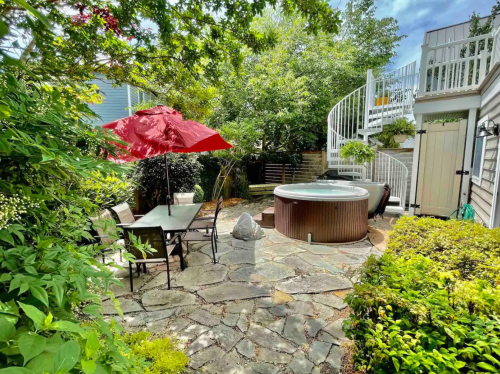 A serene backyard with a hot tub, dining table under a red umbrella, and lush greenery surrounding a stone patio.