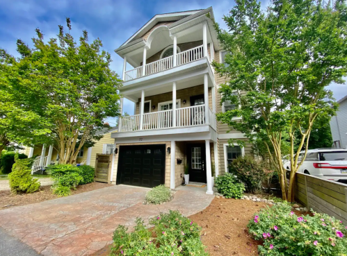 A three-story house with balconies, surrounded by greenery and flowers, featuring a stone driveway and a black garage door.