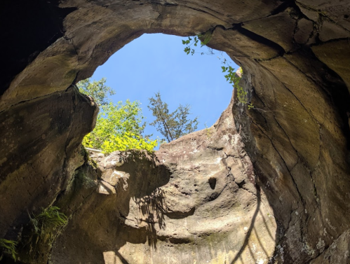 View looking up from inside a rocky cave, framed by stone walls and greenery against a bright blue sky.