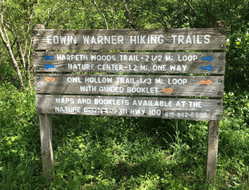 Wooden sign for Edwin Warner Hiking Trails, listing trail names, distances, and directions in a green, wooded area.
