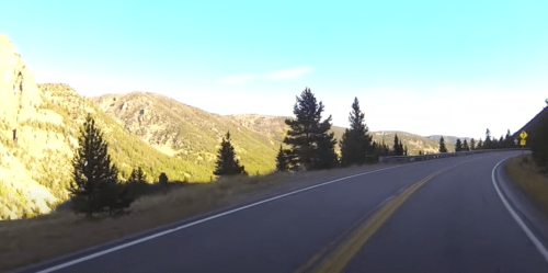 Curving mountain road with trees and hills under a clear blue sky.