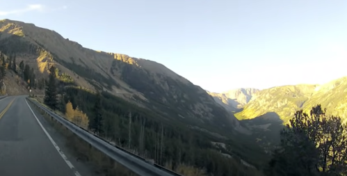 Scenic mountain road with sunlit peaks and valleys, surrounded by trees and rocky terrain.