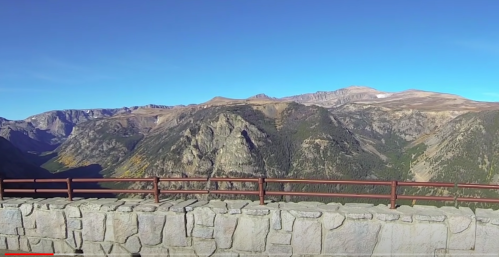 A panoramic view of mountains and valleys under a clear blue sky, with a stone railing in the foreground.