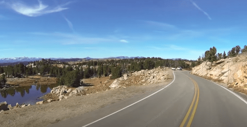 A winding road through a scenic landscape with trees, rocks, and distant mountains under a clear blue sky.