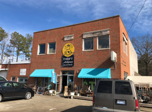 A brick thrift store with blue awnings, featuring a sign that reads "Thrift Antiques Collectibles" and parked vehicles outside.