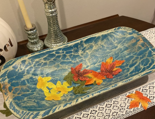 A blue decorative tray with colorful autumn leaves on a lace table runner, alongside a candle and silver candlestick.