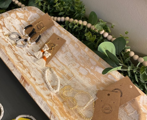 A wooden tray displays various handmade jewelry pieces, including earrings and bracelets, surrounded by greenery.