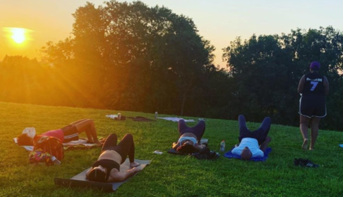 People practicing yoga on a grassy field at sunset, with a warm glow and trees in the background.