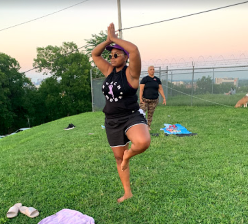 A person practicing yoga on one leg in a grassy area, with another person in the background.