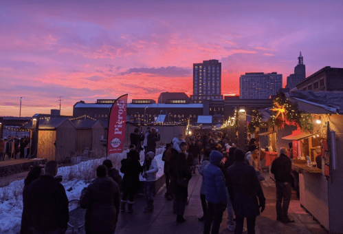 A vibrant sunset casts pink and purple hues over a bustling market with festive lights and people enjoying the scene.