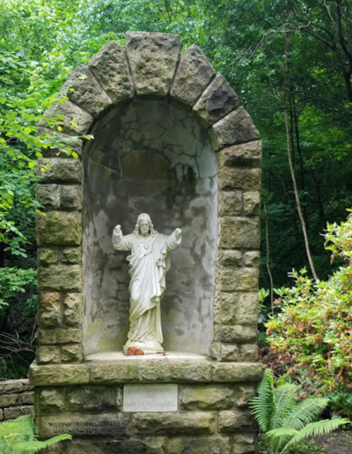 A stone archway with a statue of Jesus surrounded by greenery in a serene outdoor setting.