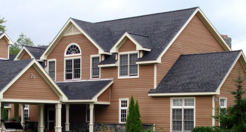 A large, two-story house with brown siding, white trim, and a gabled roof, surrounded by greenery.