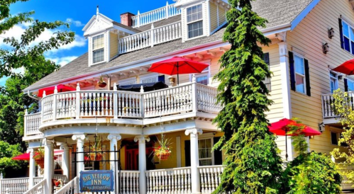 A charming yellow inn with a wraparound porch, red umbrellas, and lush greenery under a bright blue sky.