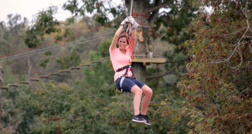 A woman in a pink shirt and shorts zip-lining through a forested area, smiling as she enjoys the adventure.