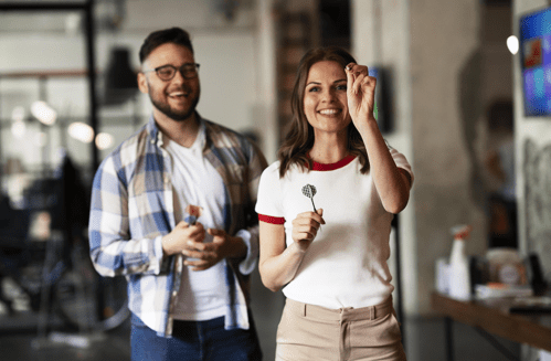 A smiling woman holds a dart, while a man laughs in the background, both in a casual indoor setting.
