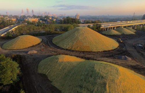 Aerial view of large, grassy mounds in an industrial area, with a city skyline in the background and morning fog.