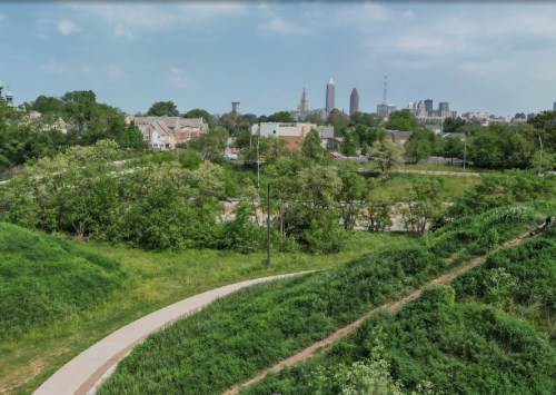 A green landscape with a winding path, featuring trees and buildings of a city skyline in the background.