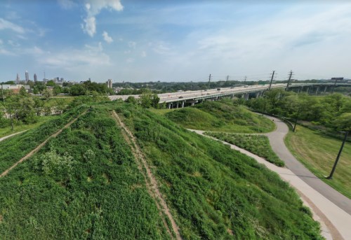 A green hillside overlooks a highway, with city buildings in the background under a clear blue sky.