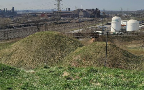 Two grassy mounds in the foreground with industrial buildings and storage tanks in the background under a clear sky.