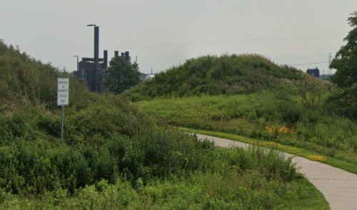 A grassy path leads to a hill, with industrial buildings visible in the background and a warning sign nearby.
