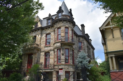 Victorian-style stone house with red accents, surrounded by greenery and a wrought-iron fence under a cloudy sky.