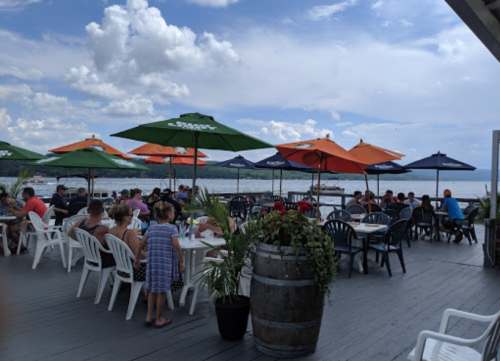 Outdoor dining area with people seated under colorful umbrellas, overlooking a body of water on a cloudy day.