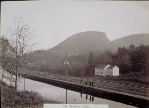 Black and white photo of a mountain with a canal and small houses, featuring four people walking along the water's edge.