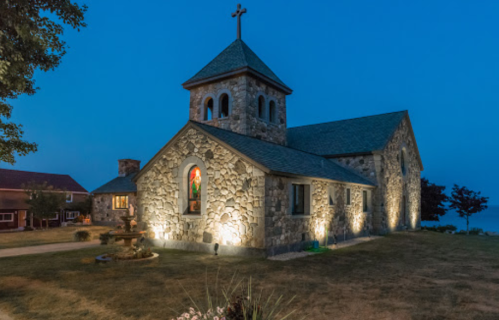 A stone church illuminated at dusk, featuring a cross on the steeple and a colorful stained glass window.