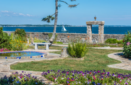 A scenic garden with colorful flowers, a stone wall, and a sailboat on the water in the background under a clear blue sky.