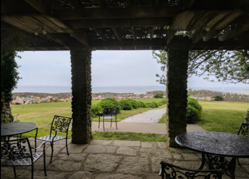 View from a stone patio with metal tables and chairs, overlooking a grassy area and distant ocean.