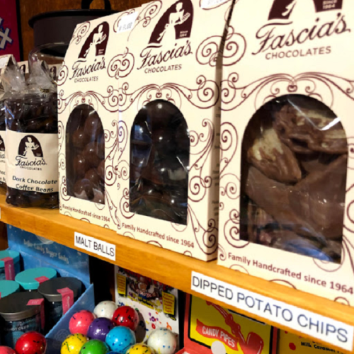A shelf displaying Fascia's Chocolates, including malt balls and dipped potato chips, with colorful candy in the background.