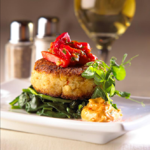 A golden crab cake topped with fresh tomatoes, served on greens with a dollop of sauce and a glass of white wine in the background.