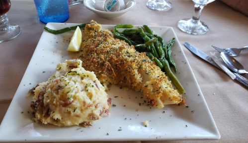 A plate with baked fish, lemon wedge, green beans, and creamy mashed potatoes garnished with herbs.