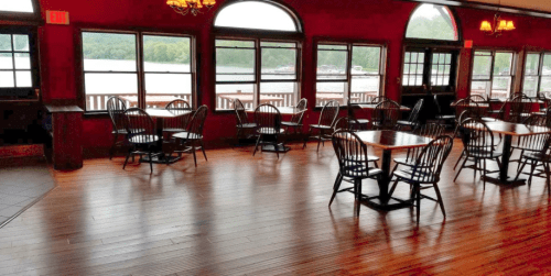 A cozy restaurant interior with wooden floors, empty tables, and large windows overlooking a lake.