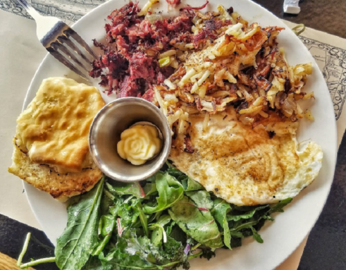 A plate of breakfast featuring eggs, hash browns, greens, and a small cup of butter.