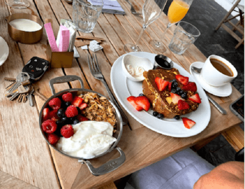 A wooden table with a breakfast spread: pancakes topped with berries, yogurt with granola, coffee, and a mimosa.