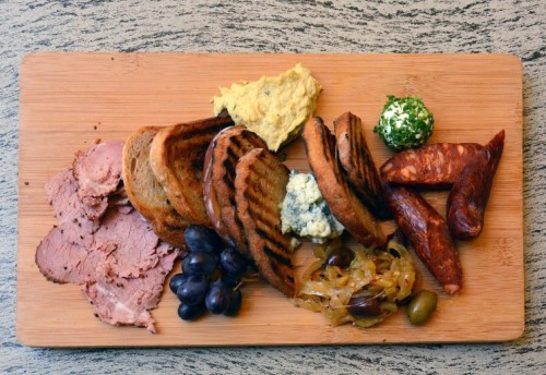 A wooden board with sliced meats, toasted bread, olives, and dips, arranged artfully for a charcuterie platter.
