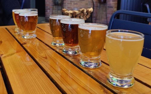 A row of six small glasses of different beers on a wooden table, with a cozy background.
