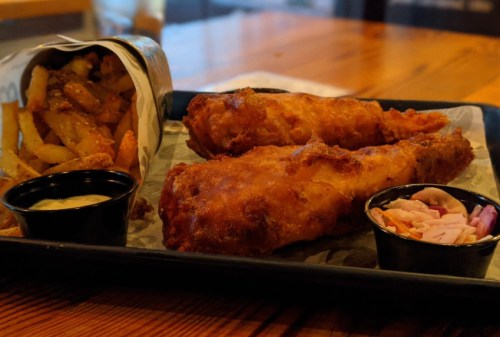 A plate with two crispy fried fish fillets, a side of fries in a paper cone, and two small cups of dipping sauce and slaw.